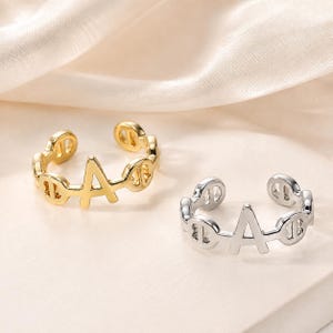 Könnte beinhalten: Zwei offene Ringe, einer in Goldoptik und einer in Silberoptik, die jeweils einen zentralen Buchstaben A flankiert von kreisförmigen Gliedermotiven zeigen. Die Ringe haben eine polierte Metalloberfläche und eine verstellbare Öffnung.
