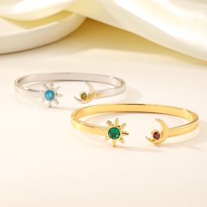 Può includere: Due bracciali aperti, uno color argento e uno color oro, ciascuno caratterizzato da un ciondolo a forma di sole con una pietra centrale e un ciondolo a forma di mezzaluna con una piccola pietra incastonata. Il bracciale argento ha sole blu e luna gialla, quello oro sole verde e luna rossa.