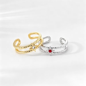 Puede incluir: Dos anillos abiertos con acabado texturizado. Un anillo es de tono dorado con un adorno central en forma de estrella con una piedra clara. El otro anillo es de tono plateado con un adorno central en forma de estrella con una piedra roja. Diseño de doble banda.