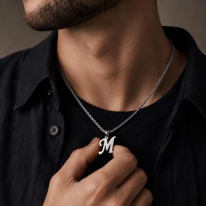 May include: A silver-toned necklace featuring a pendant shaped like the capital letter M in a cursive script font. The pendant hangs from a thin, textured rope chain that rests against a black shirt. The metal has a polished, reflective finish.