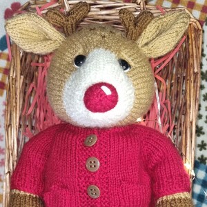 May include: A handmade knitted reindeer doll with a red sweater, green shorts, and brown hooves. The reindeer has a brown head, tan antlers, and a red nose. It is sitting in a woven basket with pink paper filler.