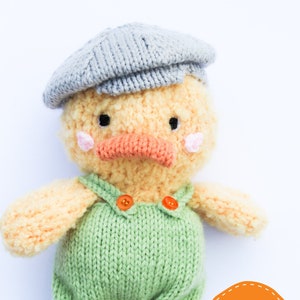 May include: A knitted yellow duck wearing a gray knitted cap and green overalls with orange buttons. The duck has a pink blush on its cheeks and an orange beak. The duck is standing on a white background. The text "CILLAS Puds" is in the lower right corner of the image.