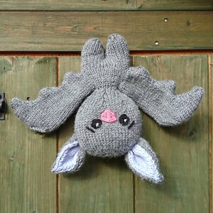 May include: A gray knitted bat with pink nose and black eyes hanging on a wooden door.