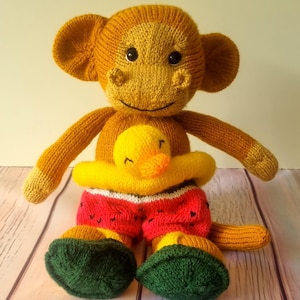 Può includere: Una bambola di scimmia lavorata a maglia a mano con un'anatra gialla attaccata. La scimmia è marrone con grandi orecchie, occhi neri e un viso sorridente. Indossa pantaloncini rossi con disegni di anguria e piedi verdi. L'anatra è gialla.