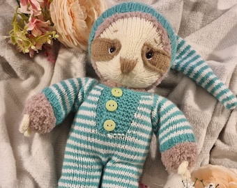 May include: A knitted sloth toy wearing a striped blue and white pajama outfit with a matching hat. The toy has brown eyes and a brown nose. The toy is lying on a white blanket.