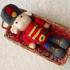 May include: A knitted nutcracker doll in a wicker basket. The doll is dressed in a red jacket with gold detailing, grey trousers, and a matching hat. The basket is lined with red paper, creating a festive holiday theme.