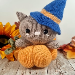 May include: A gray knitted cat wearing a blue witch hat with an orange stripe sits on top of an orange knitted pumpkin. The cat has black eyes and a white nose. The pumpkin is sitting on a white wooden surface.