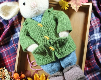 May include: A white knitted mouse wearing a green knitted jacket with yellow buttons, blue trousers, and brown knitted boots. The mouse is lying on a wooden tray with autumnal decorations.