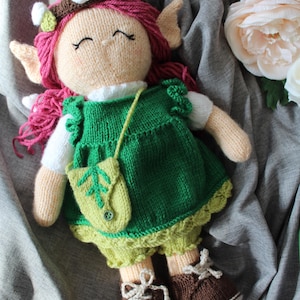 Può includere: Una bambola di lana con i capelli rosa, un vestito verde e scarpe marroni. La bambola indossa una corona di fiori e una piccola borsa verde.