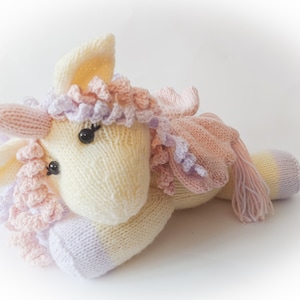 Puede incluir: Un juguete de peluche de unicornio tejido a mano con una melena y cola rosa y morada. El unicornio está acostado con la cabeza apoyada en sus patas delanteras.