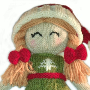May include: Knit doll with a white and red Santa hat, a green sweater with a white snowflake design, and red bows in her hair.