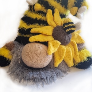 May include: A knitted gnome wearing a yellow and black striped hat with a fuzzy texture. The hat has a large yellow sunflower with a brown centre and three small bees with white wings.