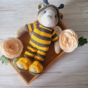 May include: A knitted bee-themed teddy bear wearing a yellow and black striped outfit and a black and gray hat. The bear is lying on a wooden tray with two peach-colored flowers.