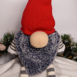 May include: A red knitted gnome with a gray fur body and gray and white striped legs. The gnome is wearing a gray knitted hat and is sitting on a white fluffy surface.