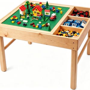 May include: A light-colored wooden activity table featuring a green building block baseplate on one side with various colorful plastic house and vehicle structures, and three storage compartments on the other side filled with wheels and blocks.