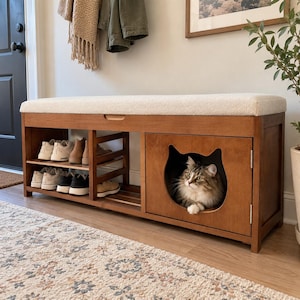 May include: A wooden entryway bench with a cream-colored upholstered seat, featuring open shelving for shoes on the left and a closed cabinet on the right with a cat-shaped cutout entrance containing a long-haired brown and white tabby cat.