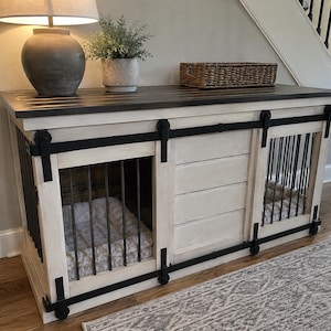 May include: A white wooden dog crate console with a dark brown top, featuring two sliding barn-style doors with black metal hardware and vertical bars. The unit includes a central storage section and two interior pet beds with light-colored cushions.