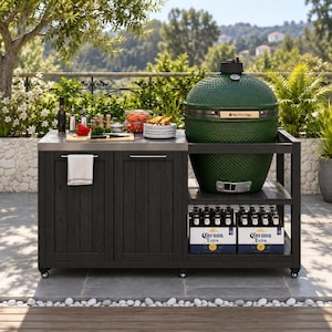 May include: A dark charcoal outdoor kitchen island with a stainless steel countertop and a large, textured, forest green ceramic kamado-style grill. The unit features two lower cabinets, a side shelf holding two cases of Corona Extra beer, and wheels.