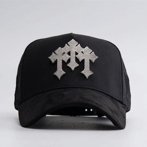 Puede incluir: Una gorra de béisbol negra con un prominente emblema de tono plateado en el panel frontal. El emblema consta de tres formas de cruz interconectadas con un acabado metálico texturizado. La gorra tiene una corona estructurada y una visera curva.