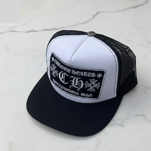 May include: A black and white trucker hat featuring a white front panel with a black rectangular patch. The patch displays the text CHROME HEARTS, the letters CH, and the text HOLLYWOOD, USA, accompanied by cross symbols in a gothic font style.
