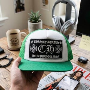 May include: A green and white trucker hat featuring a rectangular black patch on the front panel. The patch displays the text CHROME HEARTS, the letters CH, and HOLLYWOOD, USA, accompanied by two cross symbols in a gothic font style.