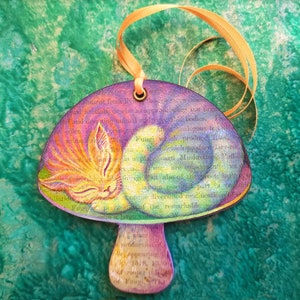 Può includere: Un ornamento a forma di fungo colorato con un gatto che dorme sopra. Il fungo è dipinto con un arcobaleno di colori e ha un motivo testuale sulla superficie. Il gatto è arancione e bianco con una coda soffice.