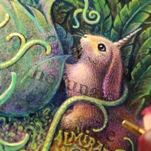May include: A close-up of a pink and purple rabbit with a single horn, surrounded by green leaves and vines. The rabbit is looking up and has a curious expression. The word "ALMIRAS" is visible in the lower right corner of the image.