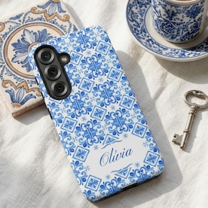 Può includere: Una custodia rettangolare per smartphone caratterizzata da un motivo a piastrelle blu e bianco con un disegno floreale ripetuto. La parte inferiore presenta un'area rettangolare bianca con il nome Olivia stampato in corsivo blu.