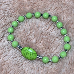 May include: A stretch bracelet featuring round, smooth, light green beads alternating with small, metallic silver-toned spacer beads. A single, larger, oval-shaped green bead with a dark green crackle pattern is centered between two silver-toned metal tubes.