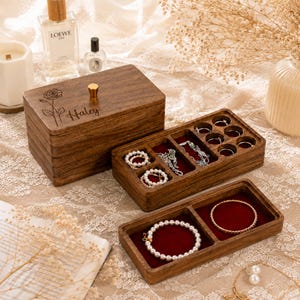 May include: A dark brown wooden jewelry box with a stacked design, featuring a gold knob and a laser-engraved rose and the name Haley on the lid. The box includes multiple open compartments lined with deep red velvet, holding various rings and bracelets.