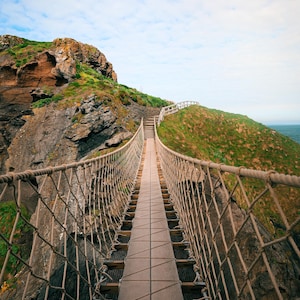 May include: A rope suspension bridge stretches between two green, grassy hills and a rocky cliff face under a cloudy blue sky. A bird flies overhead. The bridge has wooden planks and rope netting.