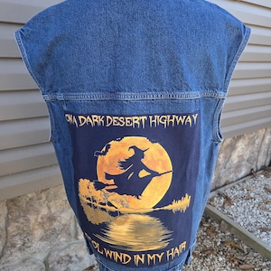 May include: A sleeveless denim jacket with a dark blue patch featuring a witch silhouette flying in front of a full moon. The patch includes the text "ON A DARK DESERT HIGHWAY" and "WIND IN MY HAIR".