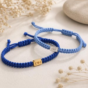 Op de afbeelding: Twee verstelbare armbanden van gevlochten koord. De ene is donkerblauw met een goudkleurige rechthoekige kraal gegraveerd met de letters K, J en een hartje. De andere is lichtblauw met een zilverkleurige kraal gegraveerd met de letters S en E.