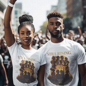 Op de afbeelding: Twee mensen dragen witte t-shirts met de tekst "STILL RISING" en een afbeelding van silhouetten. De vrouw heft haar vuist. De shirts bevatten ook extra tekst en een afbeelding van mensen.