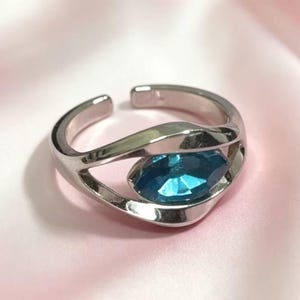 May include: A silver-toned open ring featuring a polished, fluid band that curves around a single, marquise-cut bright blue gemstone. The metal band has a smooth, reflective finish and an adjustable gap at the back, showcasing a sleek design.