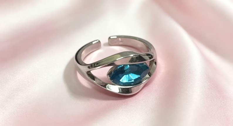 May include: A silver-toned open ring featuring a polished, fluid band that curves around a single, marquise-cut bright blue gemstone. The metal band has a smooth, reflective finish and an adjustable gap at the back, showcasing a sleek design.