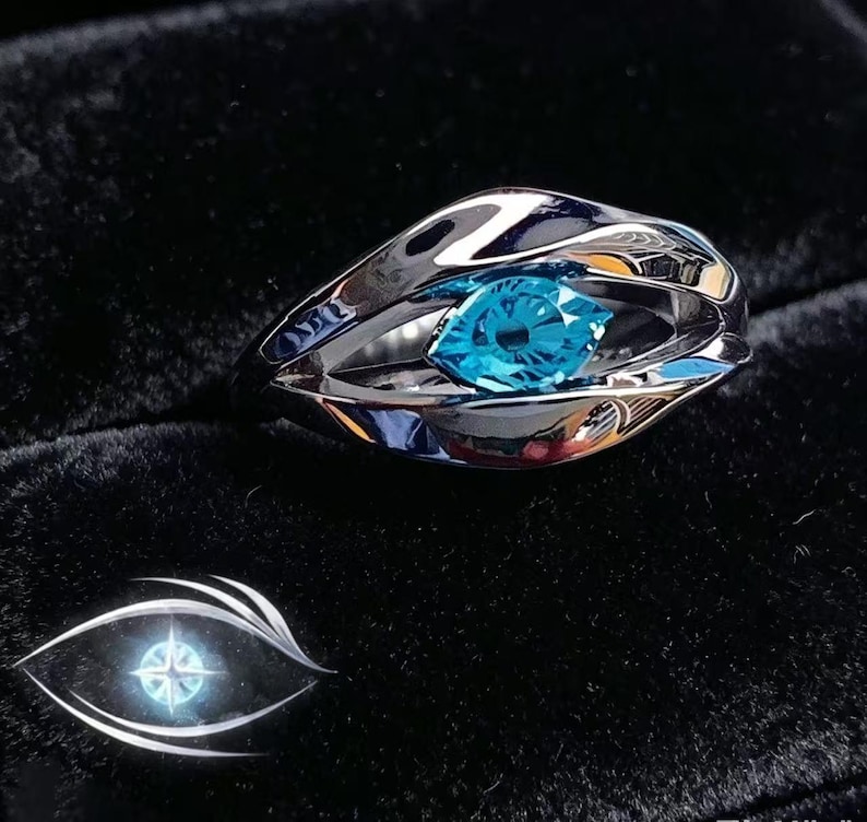 May include: A silver-toned ring featuring a polished, fluid metal band that curves to form an eye-shaped setting. A single, brilliant, bright blue gemstone is securely held in the centre of the metal frame, creating a striking contrast.