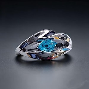 May include: A polished silver-tone ring featuring a central marquise-cut bright blue gemstone. The metal band has a fluid, overlapping design that curves around the stone, creating a layered appearance with reflective surfaces and smooth edges.