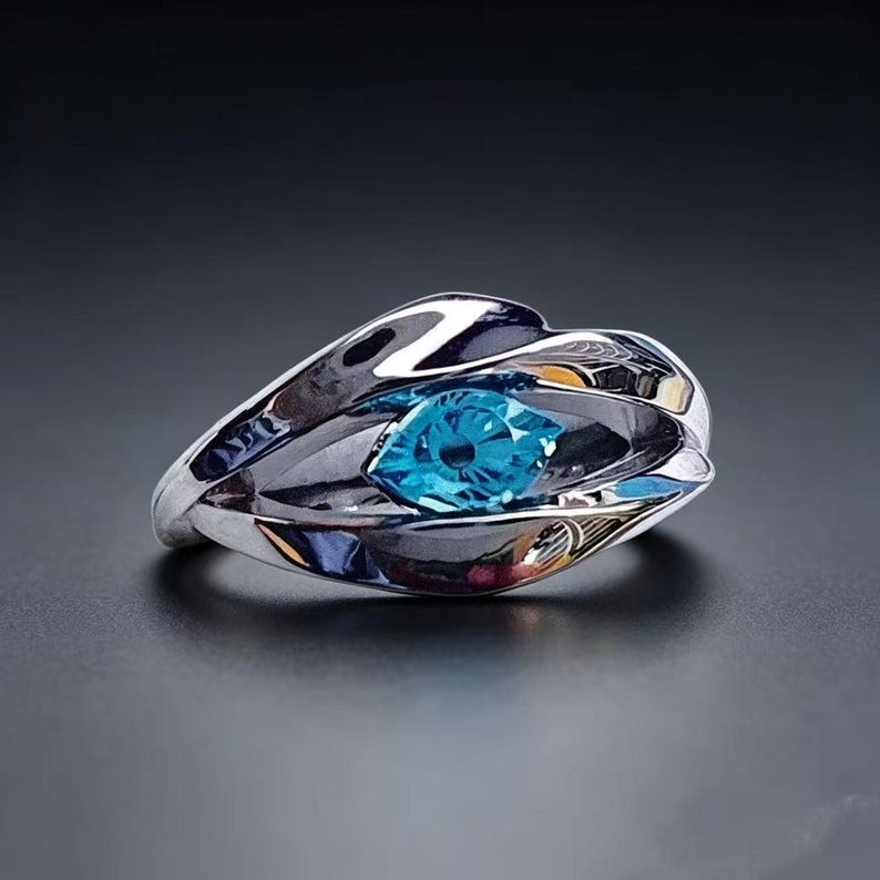 May include: A polished silver-tone ring featuring a central marquise-cut bright blue gemstone. The metal band has a fluid, overlapping design that curves around the stone, creating a layered appearance with reflective surfaces and smooth edges.