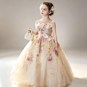 May include: A floor-length beige tulle dress featuring a sheer illusion neckline and intricate floral embroidery with three-dimensional fabric flowers in shades of pink, blue, and yellow, accented with sequins and a small matching beige handbag.