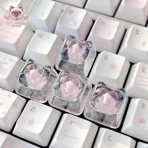 May include: Four clear, square mechanical keyboard keycaps featuring a translucent pink heart shape inside each. A small, opaque white cross is centred on the surface of each pink heart. The keycaps are positioned on a white keyboard with pink accents.