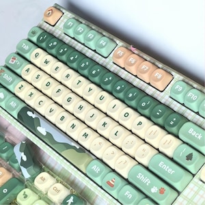 Könnte beinhalten: Eine mechanische Tastatur mit einem hellgrünen karierten Rahmen und einer Mischung aus cremefarbenen, salbeigrünen und pfirsichfarbenen Tastenkappen. Die Tasten zeigen verschiedene Symbole wie einen Kalender, einen Baum und Pfotenabdrücke.