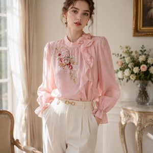 May include: A light pink long-sleeved blouse featuring a high neckline with a ruffled accent and an intricate floral embroidery detail on the chest. The blouse is paired with high-waisted cream-colored trousers that have a gold-toned metal belt buckle.