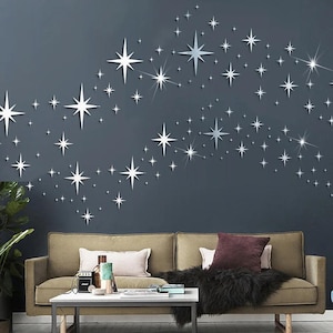 May include: A collection of silver, reflective star-shaped wall decals in various sizes arranged in a scattered pattern across a dark grey wall. The stars feature sharp, pointed rays and a metallic finish that creates a bright, celestial effect.