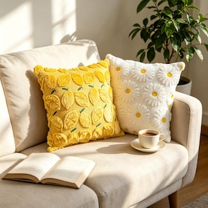 Può includere: Due cuscini quadrati, uno giallo con un motivo a limoni trapuntato e uno bianco con un motivo a margherite trapuntato, posizionati su un divano di colore chiaro accanto a un libro aperto e una tazza in ceramica bianca su un piattino.