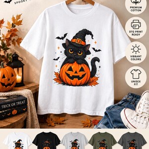 May include: A white short-sleeve t-shirt featuring a graphic of a black cat wearing a witch hat and sitting inside a carved orange jack-o'-lantern pumpkin. The design includes small black bats and orange stars. The shirt is displayed on a wooden hanger.