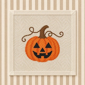 May include: A square cross-stitch embroidery piece featuring a bright orange jack-o'-lantern with a brown stem and curly vines. The pumpkin has a black triangular-eyed face and a jagged mouth, centred on a white grid fabric with a beige border.