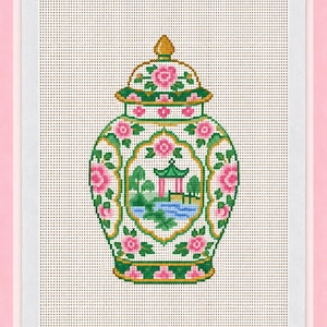 May include: A cross-stitch pattern on white fabric featuring a ginger jar with a lid. The jar is decorated with pink flowers, green leaves, and a central landscape scene depicting a pagoda, trees, and a blue body of water with gold accents.