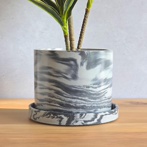 May include: A cylindrical plant pot with a matching circular saucer, both featuring a marbled pattern of swirling dark gray and white tones. The pot contains a green plant with slender stems and leaves extending upward from the center.
