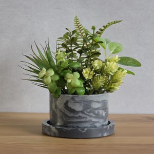 May include: A collection of various artificial green plants arranged in a cylindrical gray and white marbled planter. The planter sits on a matching circular tray, featuring a smooth finish with swirling patterns of dark and light gray tones.